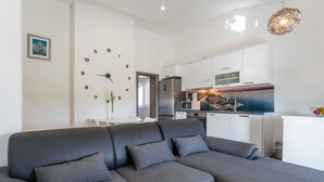 A modern living room with a grey sectional sofa, a white dining table, and a kitchen area with white cabinets and a built-in oven. - Apartments Maza (Zadar)
