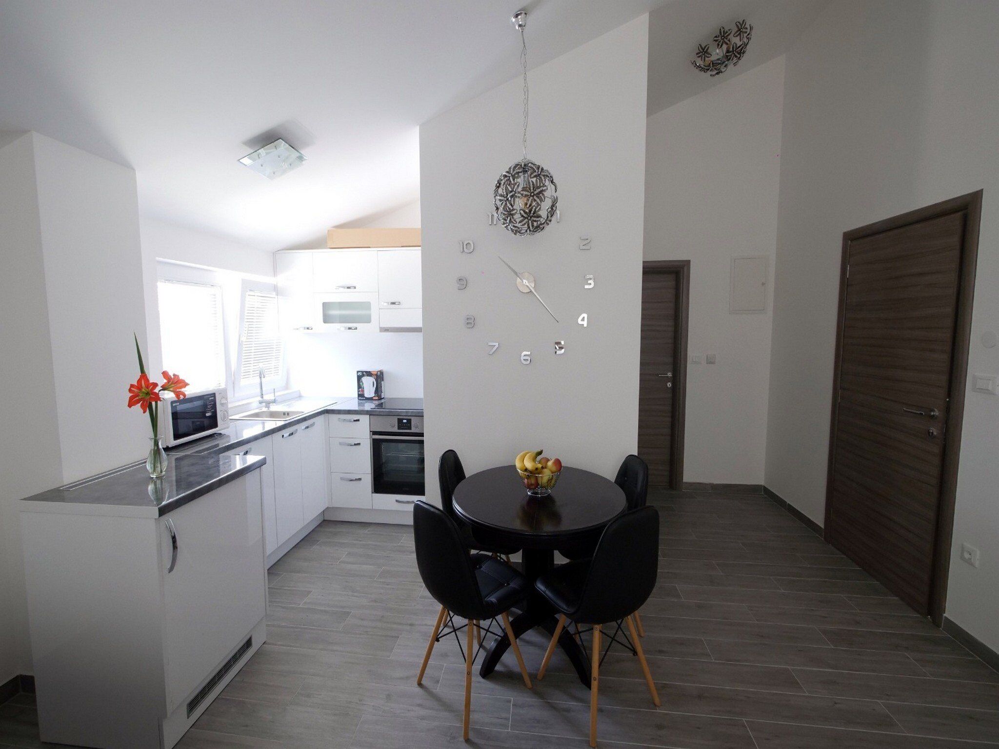 A modern kitchen with white cabinets, a microwave, and an oven. There is a dining table with two chairs and a bowl of fruit.