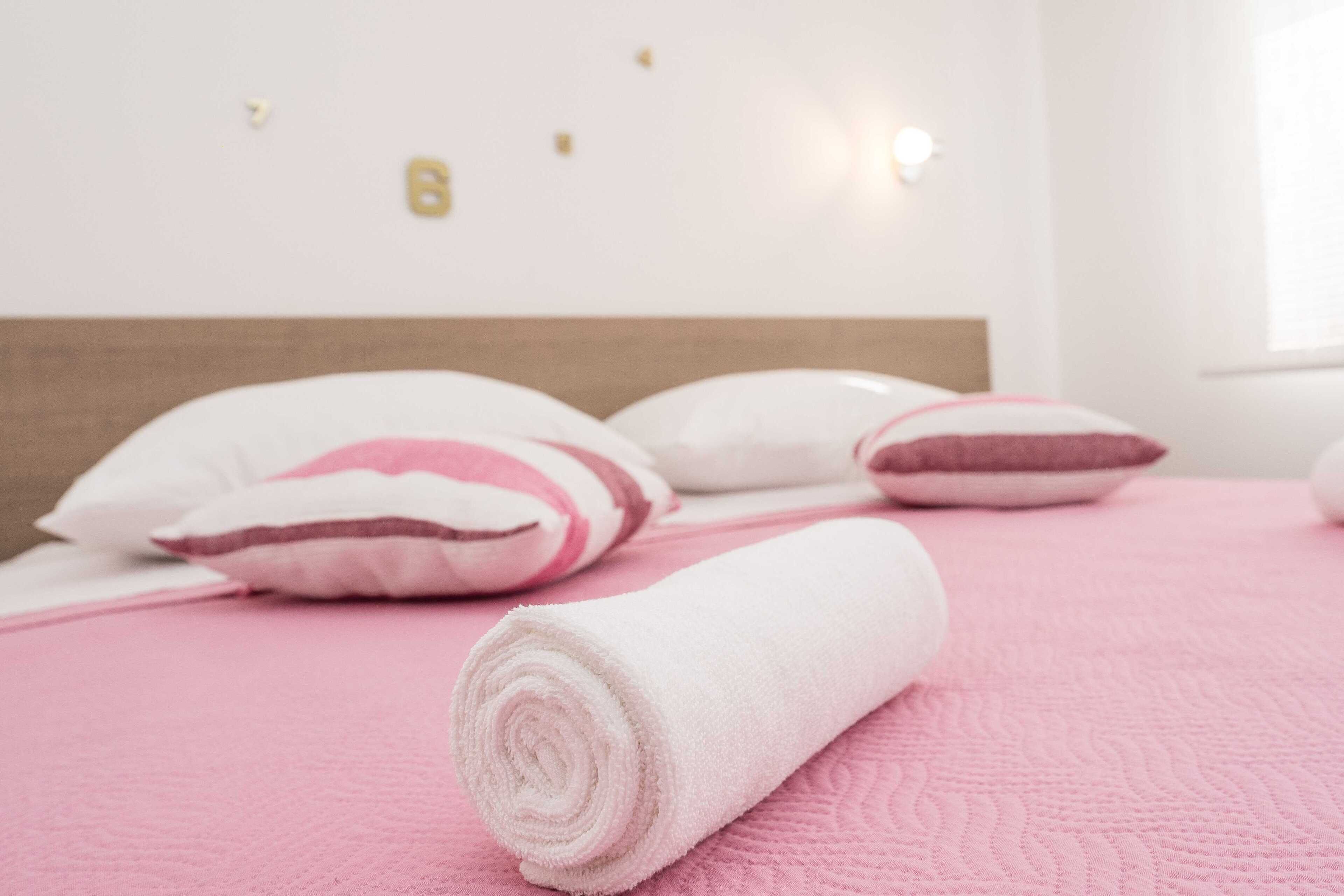 A neatly made bed with pink bedding and white pillows, a rolled white towel, and a wall with a light switch and a small decorative item.
