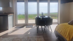 Une chambre avec une grande fenêtre, un lit recouvert d’une couverture jaune, une table à manger avec des chaises et un coin cuisine.