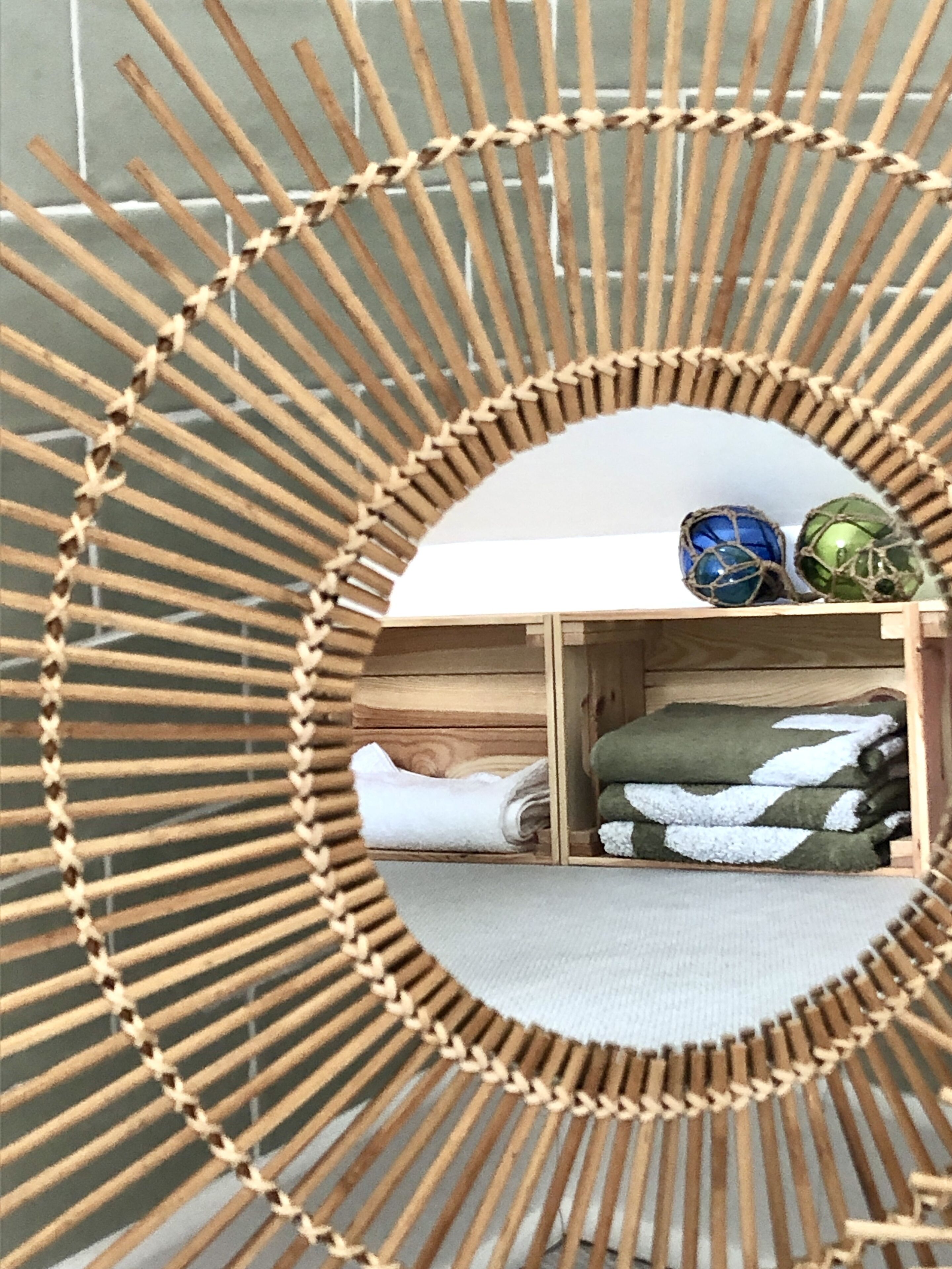 A circular woven mirror with a wooden frame, reflecting a room with a bed, a shelf with decorative items, and a folded blanket.