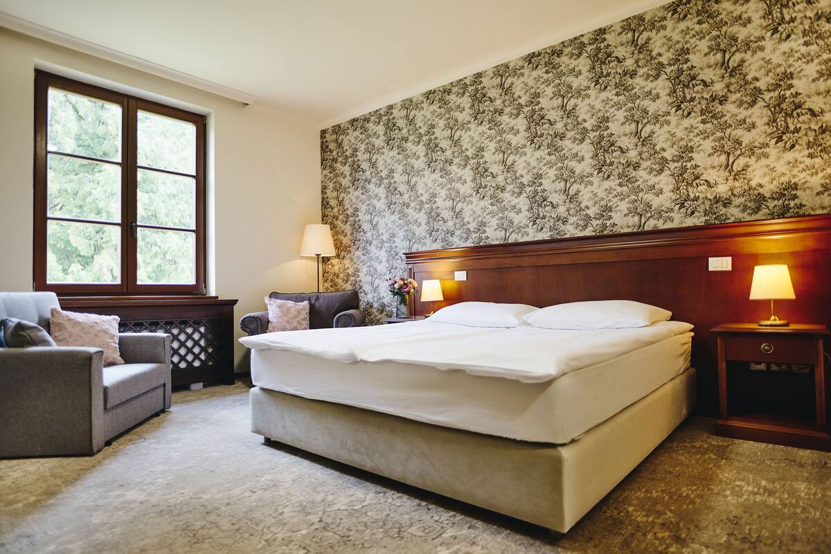 A bedroom with a large bed, a sofa, a nightstand with a lamp, and a window with a view of trees.