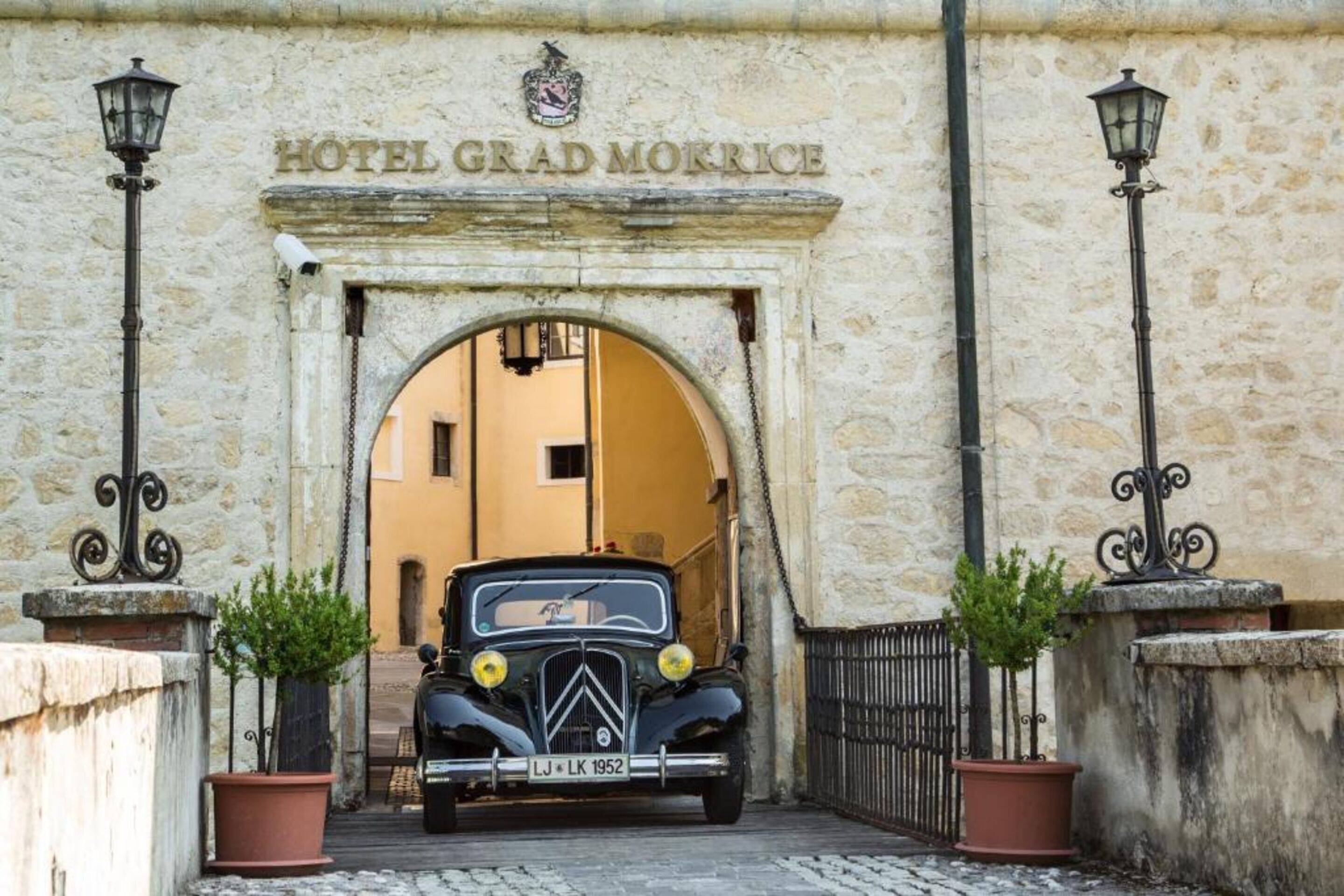 Un coche antiguo estacionado frente a la entrada del Hotel Grad Morkrice, con un arco de piedra y dos macetas a cada lado.