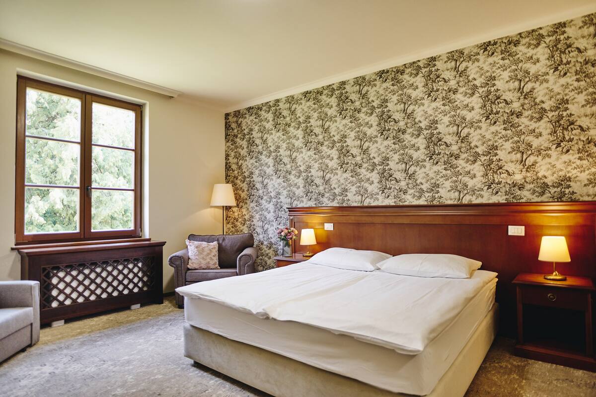 A hotel room with a large bed, two bedside lamps, a nightstand, a small table, a chair, and a window with a view of trees.