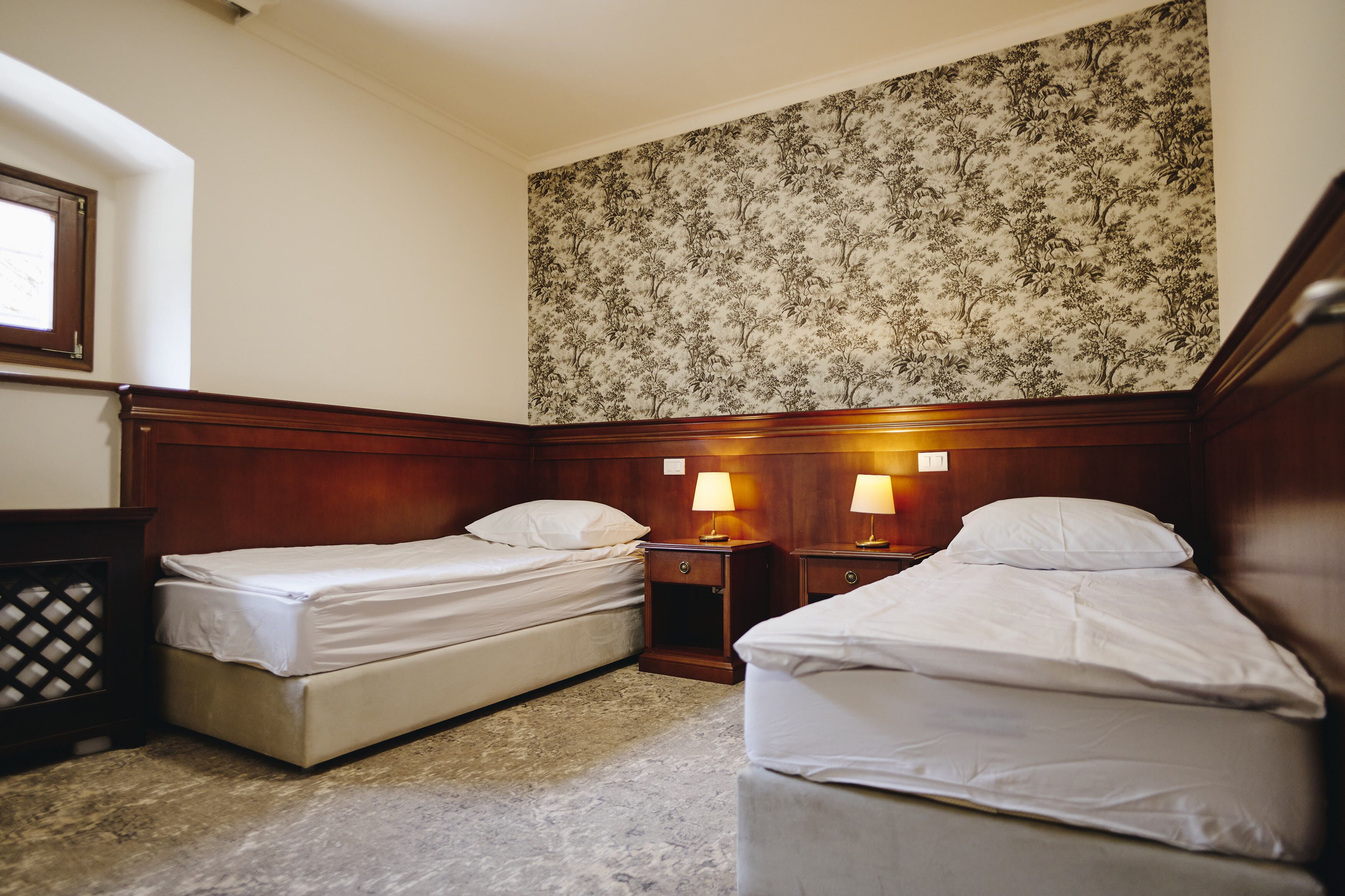 A hotel room with two single beds, wooden headboards, a floral-patterned wallpaper, and two bedside lamps.