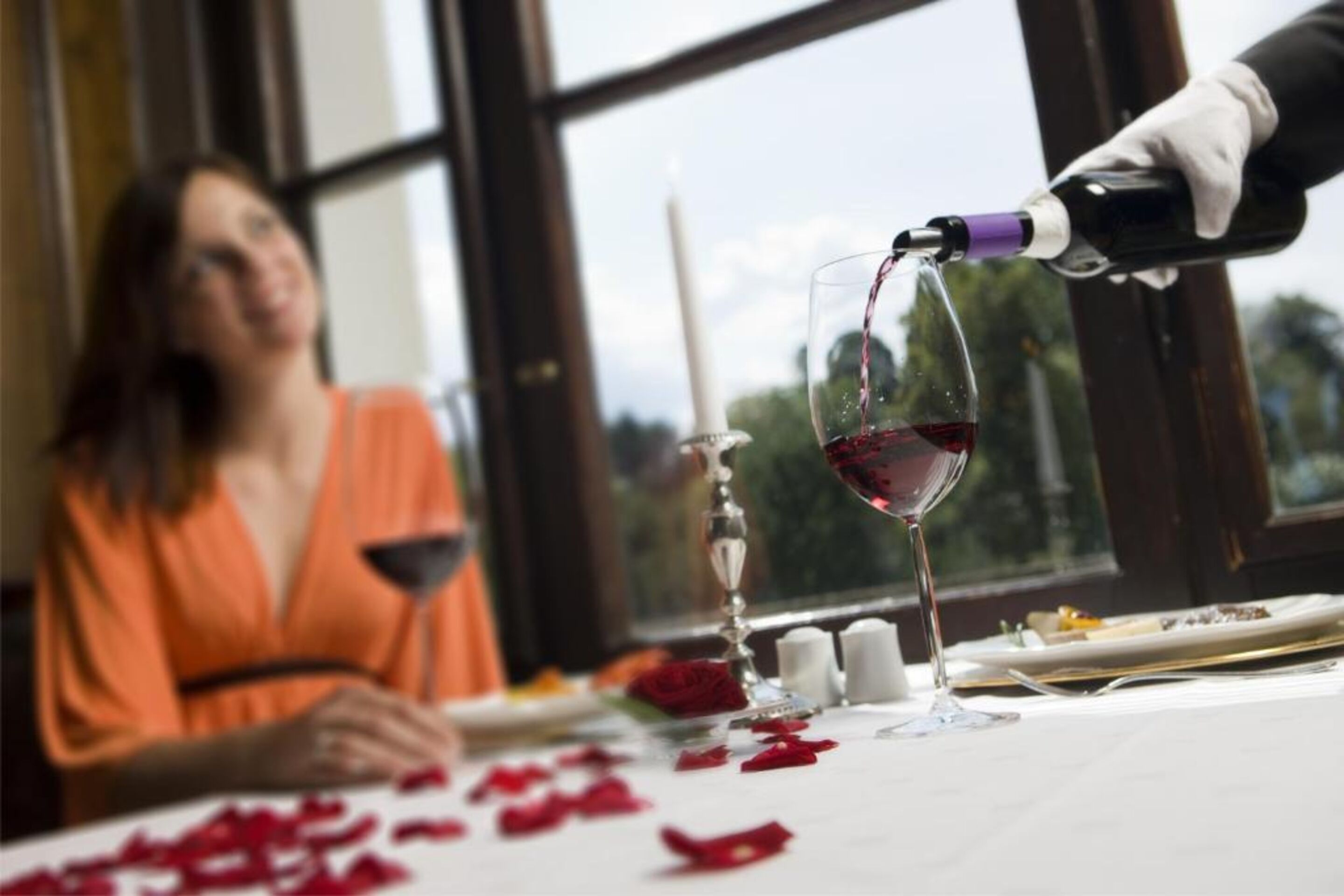 Un tavolo da pranzo con un bicchiere da vino che viene riempito, petali di rosa sparsi sul tavolo e una finestra sullo sfondo.