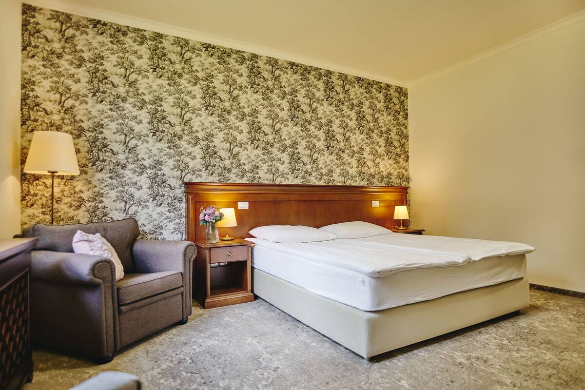 A hotel room with a bed, a chair, a nightstand with lamps, and floral wallpaper.