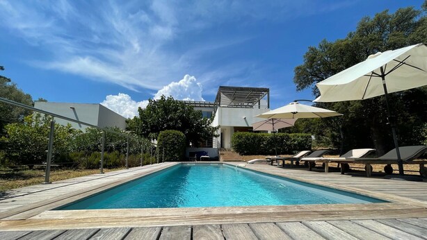 A rectangular pool with a wooden deck, surrounded by lounge chairs and umbrellas, and a modern building in the background. - Ruscana by Pinarello Villa Services (Pinarello)
