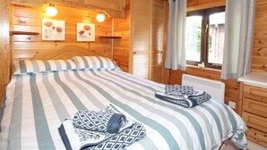 A wooden cabin bedroom with a bed, a nightstand, a window with curtains, and a wall-mounted shelf with a picture. - Robin's Nest (Keswick)