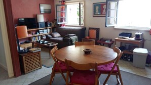 A cozy living room with a round wooden table, a sofa, a TV, a bookshelf, and a window with curtains. - Apartment (Lège-Cap-Ferret)