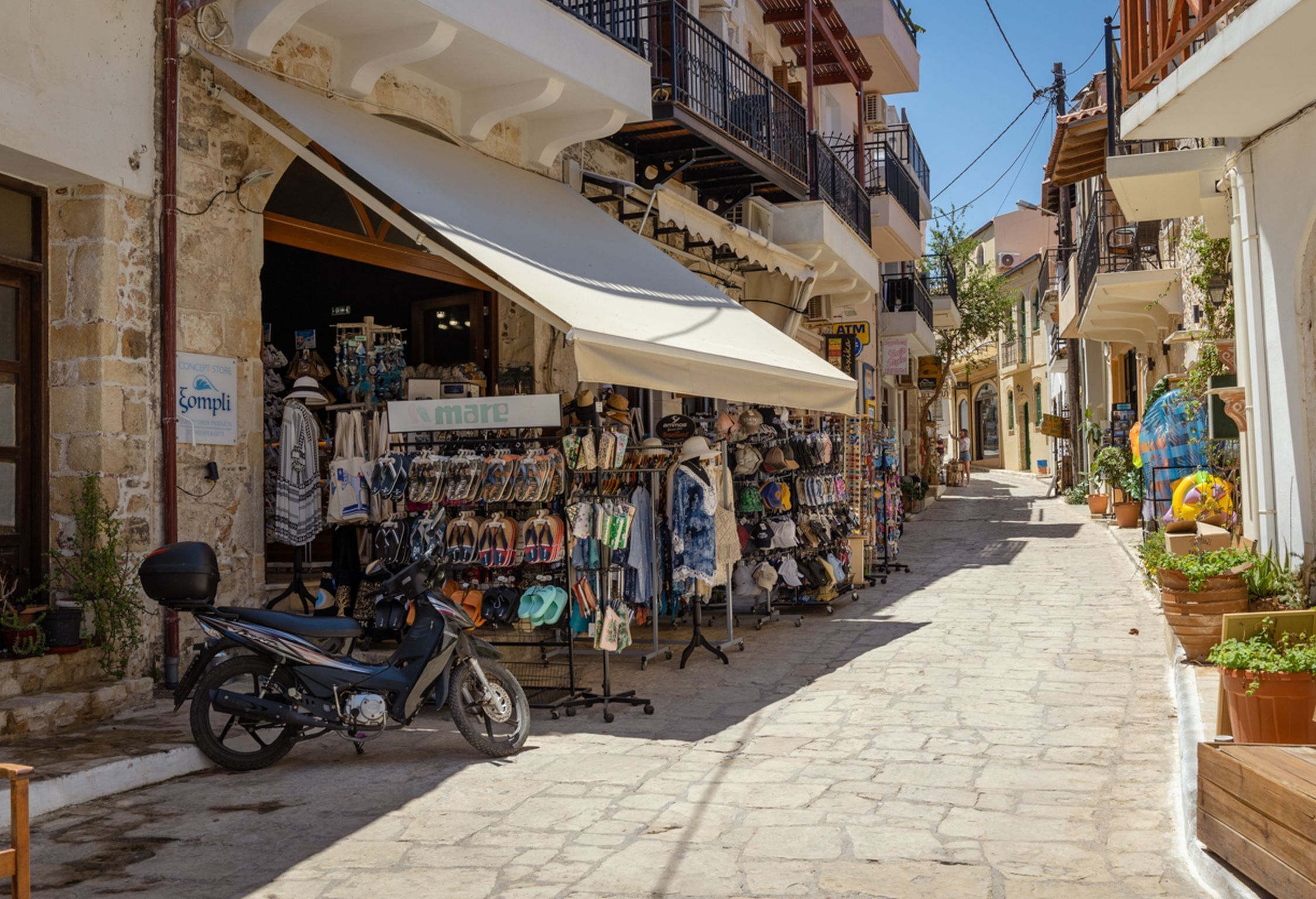 A narrow street with shops on either side, a motorcycle parked on the left, and potted plants along the right.