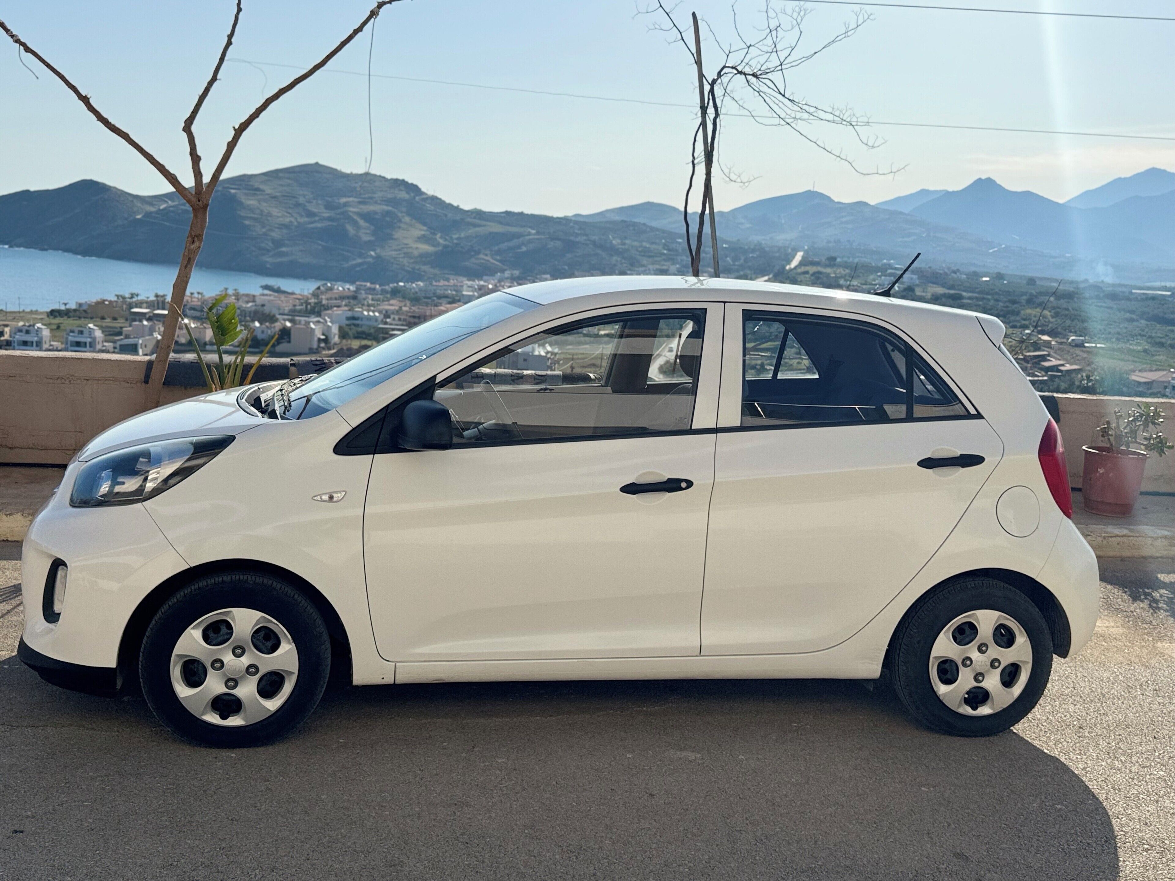 Une petite voiture blanche est garée sur une surface pavée, offrant une vue pittoresque sur les montagnes et un plan d'eau en arrière-plan.