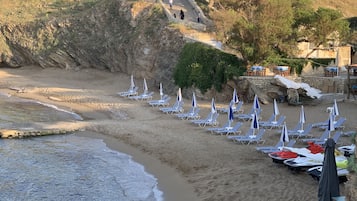 A beach with rows of sun loungers and umbrellas, a rocky cliff, and a few buildings in the background.