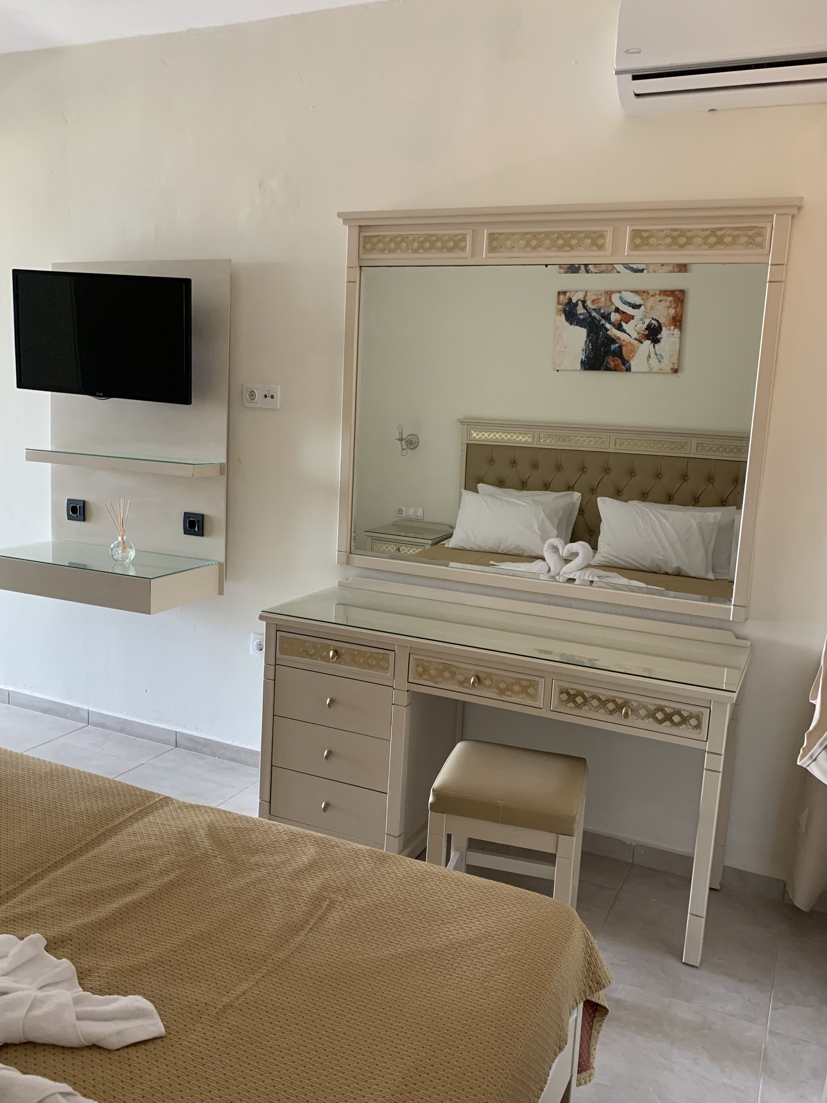 Una habitación de hotel con una cama, un tocador con espejo, un televisor montado en la pared y un cuadro sobre la cama.