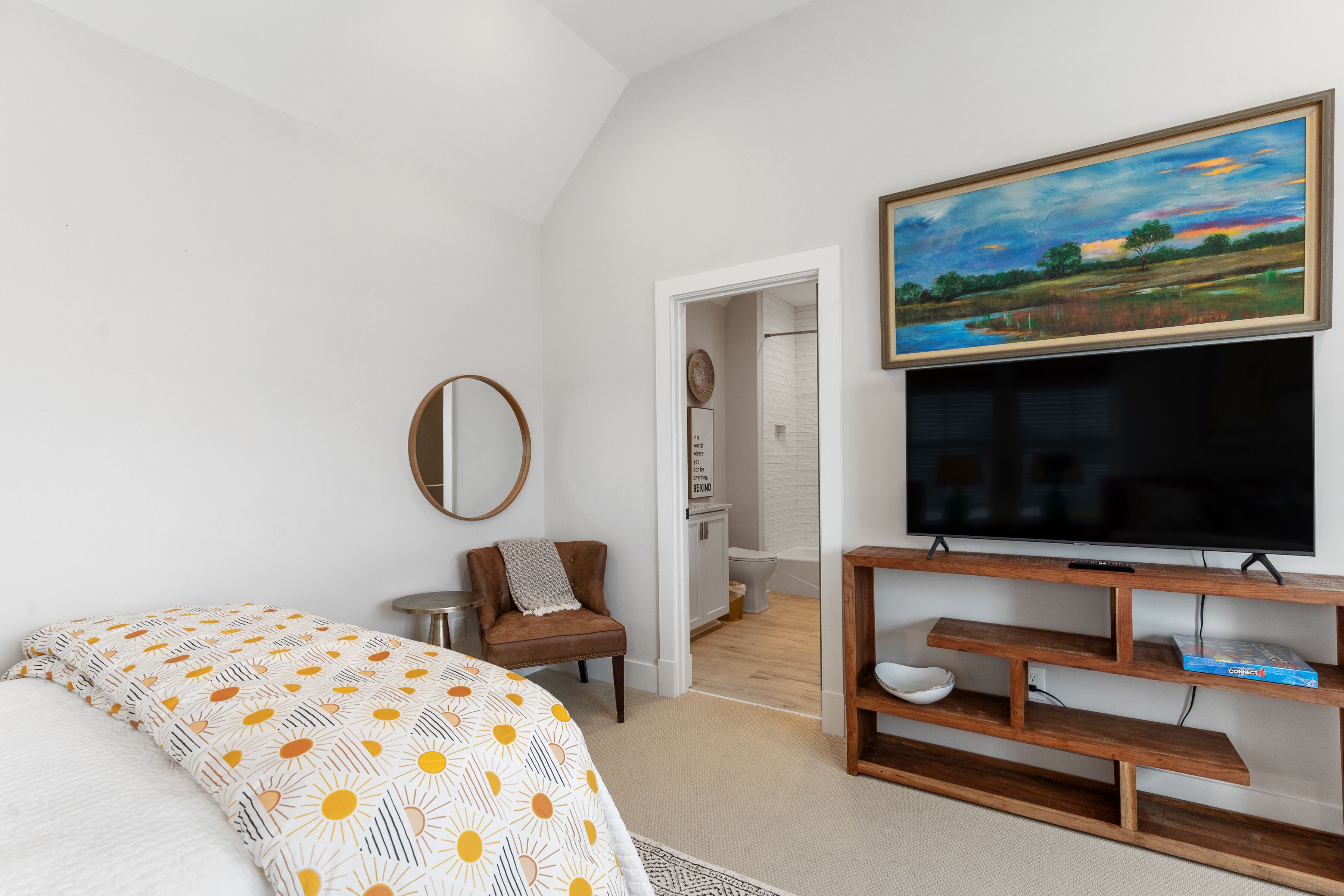 A bedroom with a bed, a TV, a painting, and a bathroom visible through an open door.