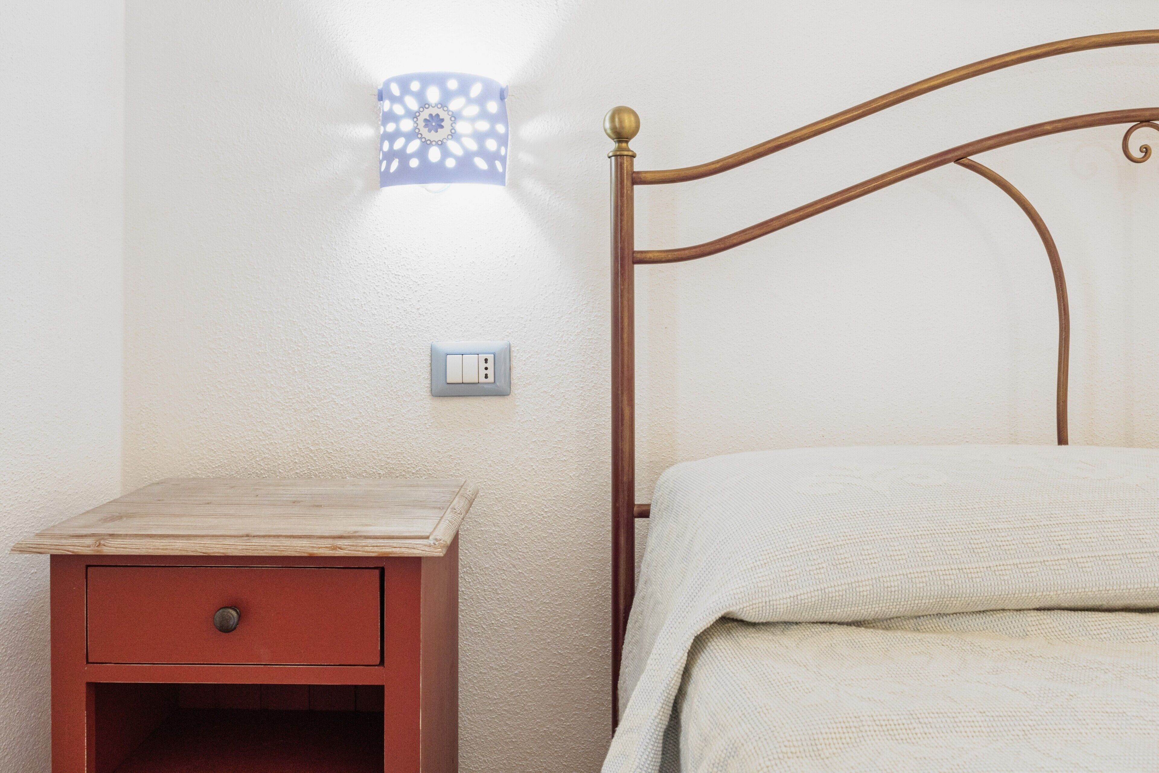A bedroom with a wooden nightstand, a bed with a white blanket, a wall-mounted light fixture, and a power outlet.