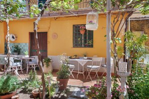 A courtyard with a yellow building, outdoor furniture, potted plants, and a hanging birdcage.