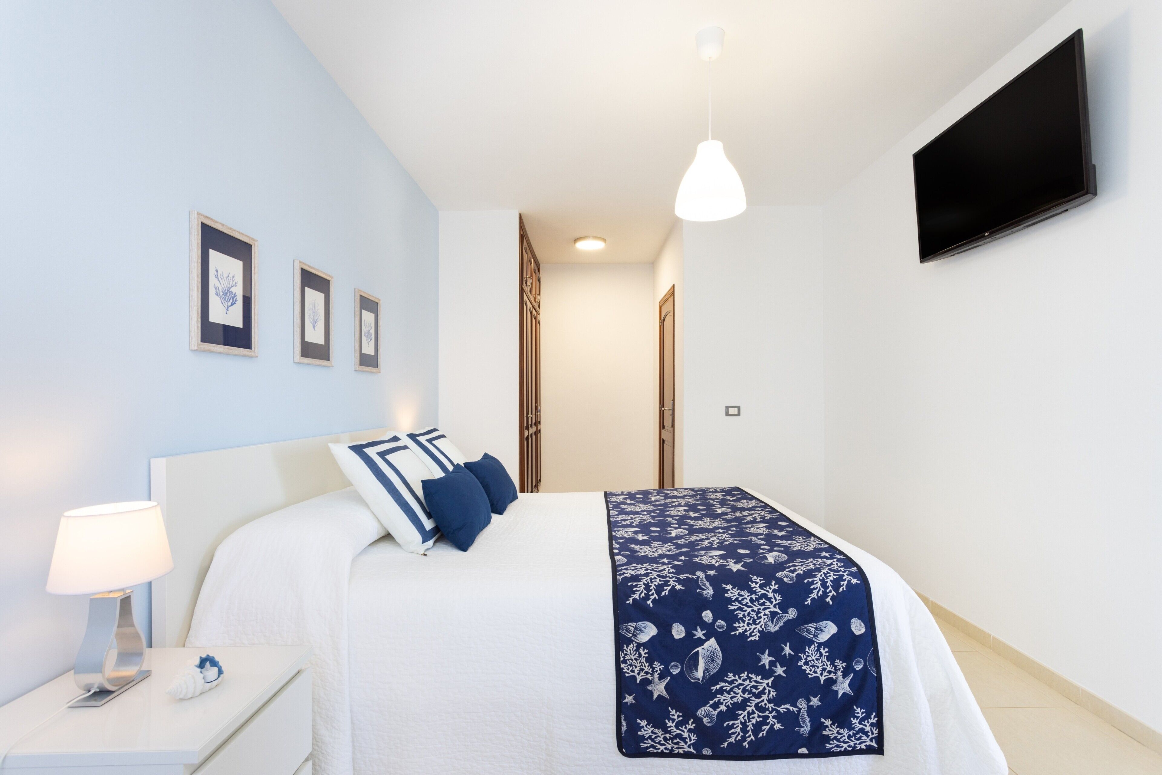 A bedroom with a bed, bedside table, lamp, wall-mounted TV, and framed pictures on the wall.