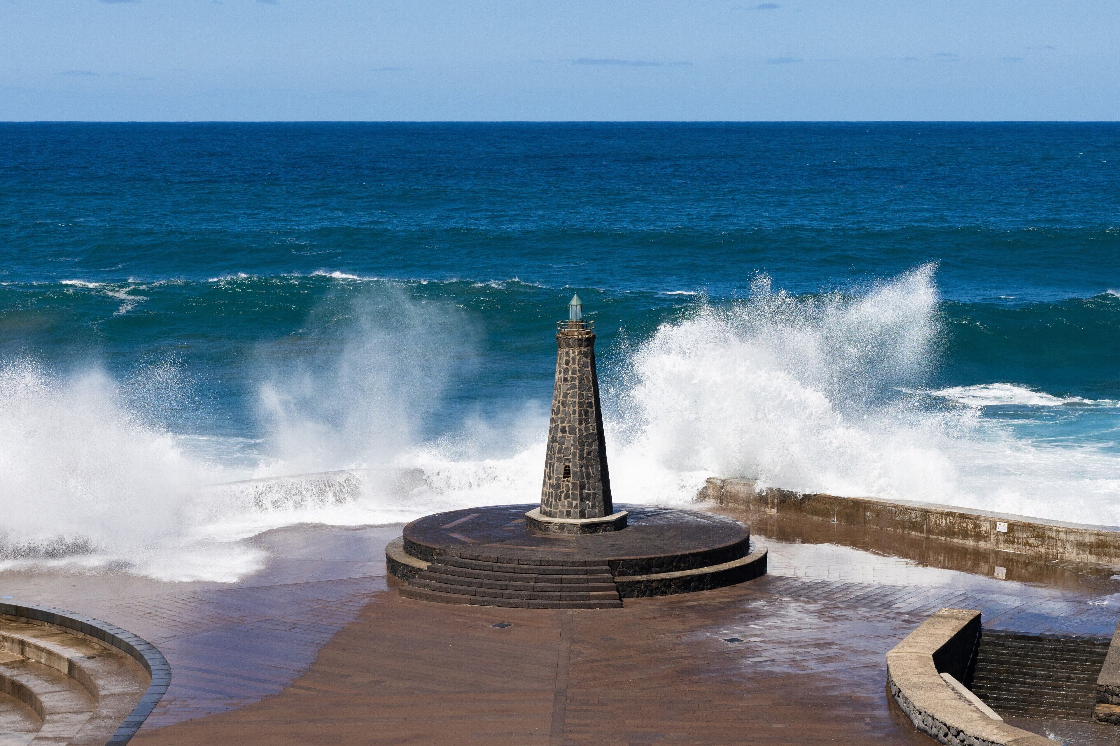 Un monument de pierre au bord de la mer, une promenade en béton et des vagues qui s’écrasent sur le rivage.
