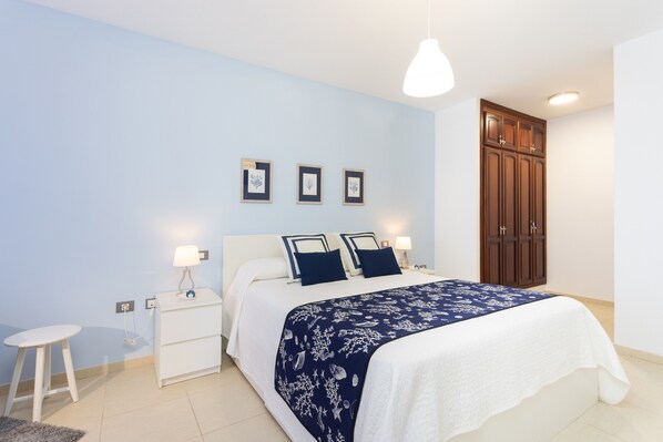 A bedroom with a bed, bedside tables, a nightstand, a lamp, a wardrobe, and framed pictures on the wall.