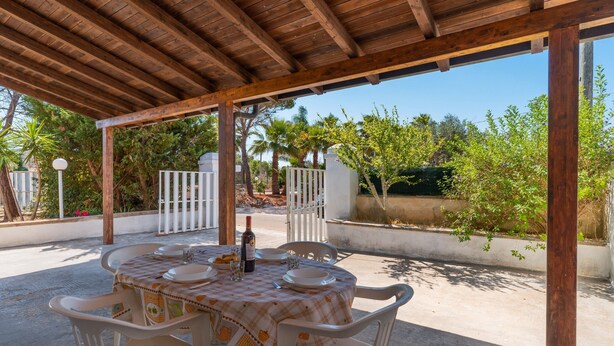 Une terrasse extérieure dotée d’une pergola en bois, d’une table dressée pour le repas et offrant une vue sur les arbres et un portail.