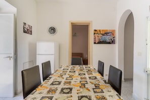 Un coin repas avec une table recouverte d’une nappe à motifs, des chaises, un réfrigérateur et une porte donnant sur une autre pièce.