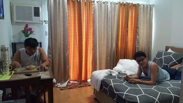 A room with a bed, a table with a cup and glasses, and a wall-mounted air conditioner. - Mesaverte Residences Afs Suites - Studio (Cagayan de Oro)
