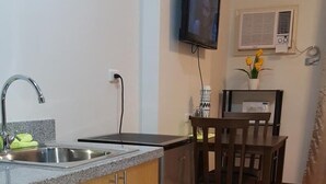 Una cocina funcional con lavamanos, un refrigerador pequeño, una mesa de comedor con sillas, un televisor montado en la pared y un equipo de aire acondicionado.