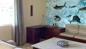 A living room with a sofa, a coffee table with books, a wooden cabinet, a wall with fish paintings, and a ceiling fan. - Beach bed and Breakfast (Saquarema)
