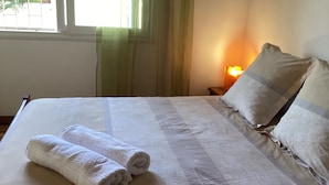 Una cama bien tendida con sábanas y almohadas blancas, una lámpara de noche y una ventana con cortinas.