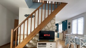 A living room with a wooden staircase, a television on a white stand, and a dining area with white chairs.