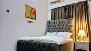 A bedroom with a tufted headboard, a bed with white bedding, a nightstand with a lamp, and an air conditioning unit on the wall. - Lovely 2-bedroom Serviced Apartment With a Balcony (Abuja)