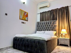 A bedroom with a tufted headboard, a bed with white bedding, a nightstand with a lamp, and an air conditioning unit on the wall.