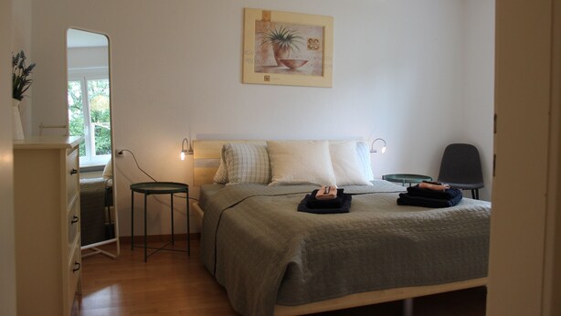 Ett sovrum med en säng, sängbord, en stol, ett skrivbord, en spegel och en hängande lampa.