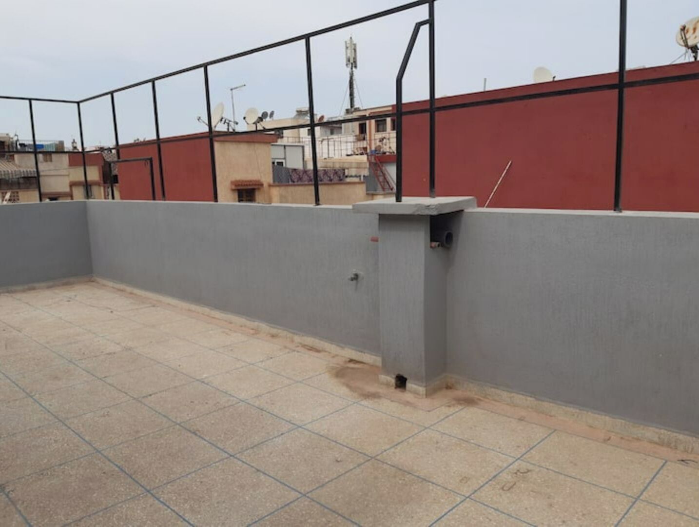 Una terraza en la azotea con suelo de baldosas, pared gris y pared roja al fondo.