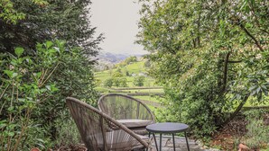 A wicker chair and table set on a stone patio with potted plants, overlooking a green landscape with hills.