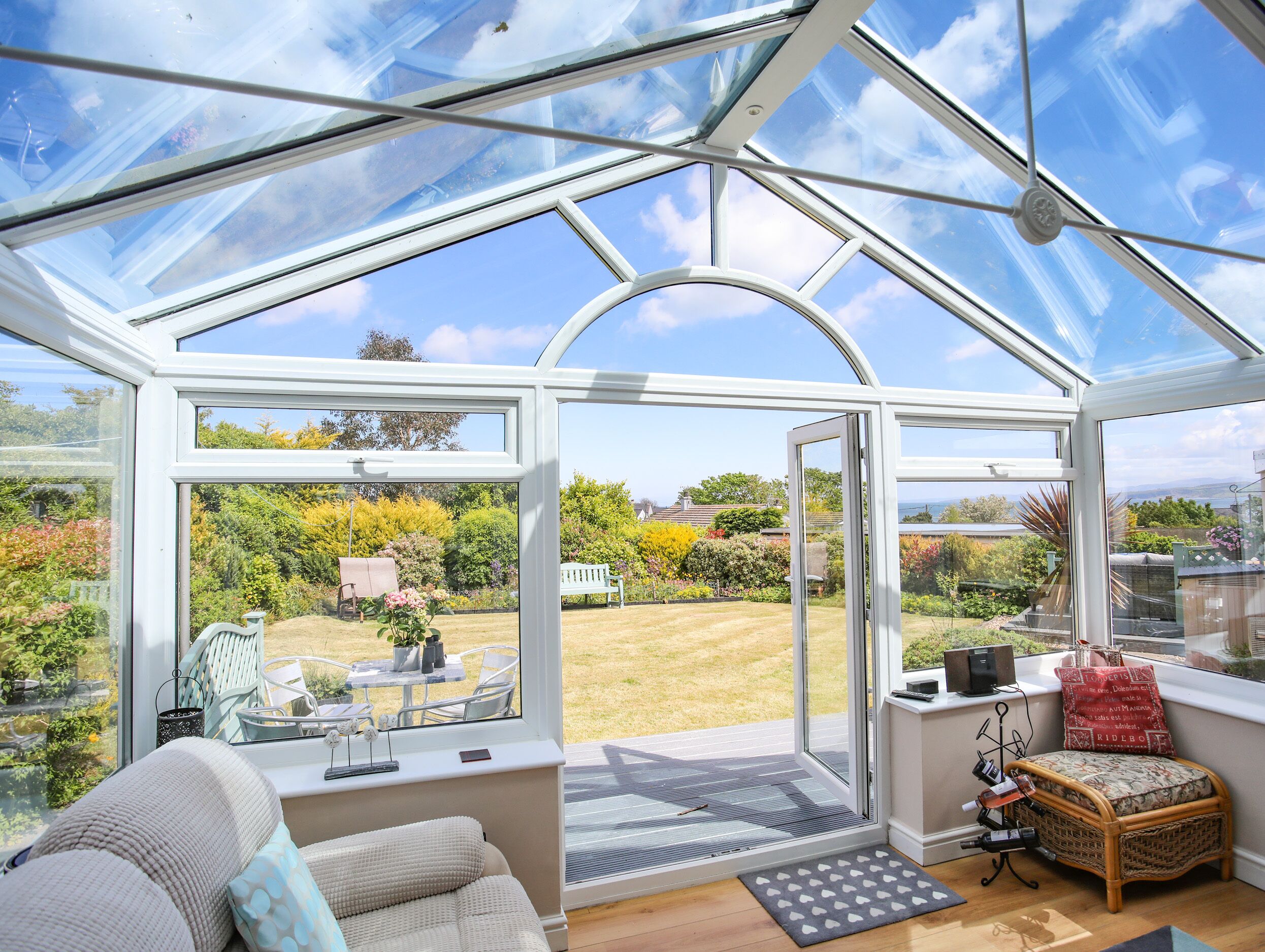 A conservatory with a glass roof, a seating area with a bench and table, and a view of a garden.