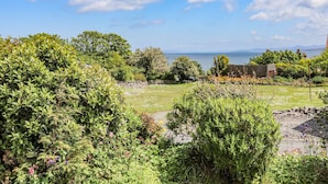Um jardim exuberante com diversas plantas e flores, um muro de pedra, e vista para o mar e montanhas ao longe.