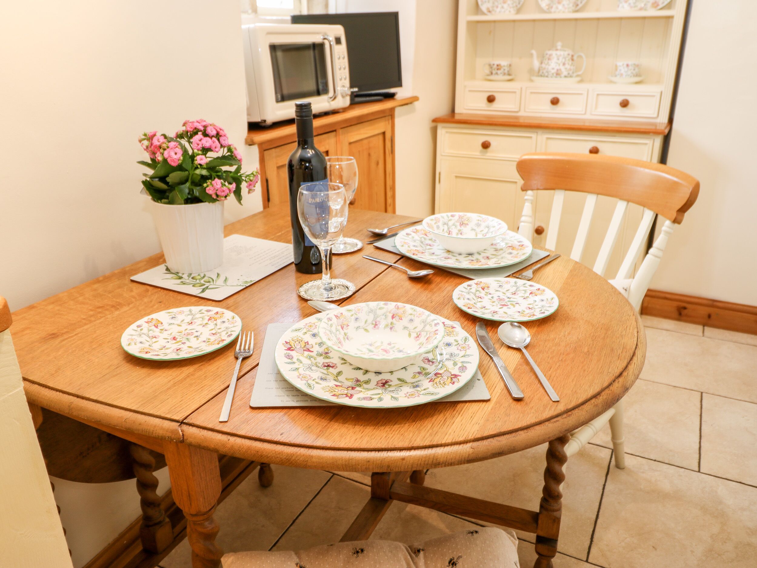 Ein Holzespeisetisch ist gedeckt mit blumengemusterten Tellern, Besteck und einer Weinflasche. Im Hintergrund sind eine Mikrowelle und Dekorationselemente zu sehen.