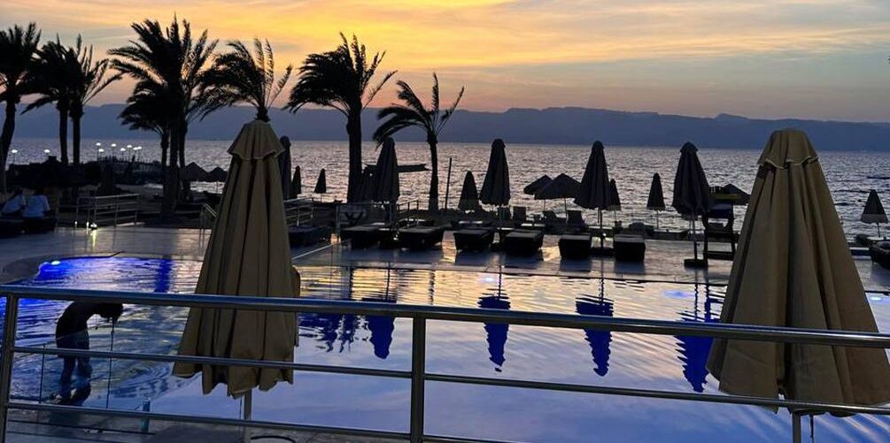 Zona de piscina con sillones y sombrillas, con vista a un cuerpo de agua al atardecer.