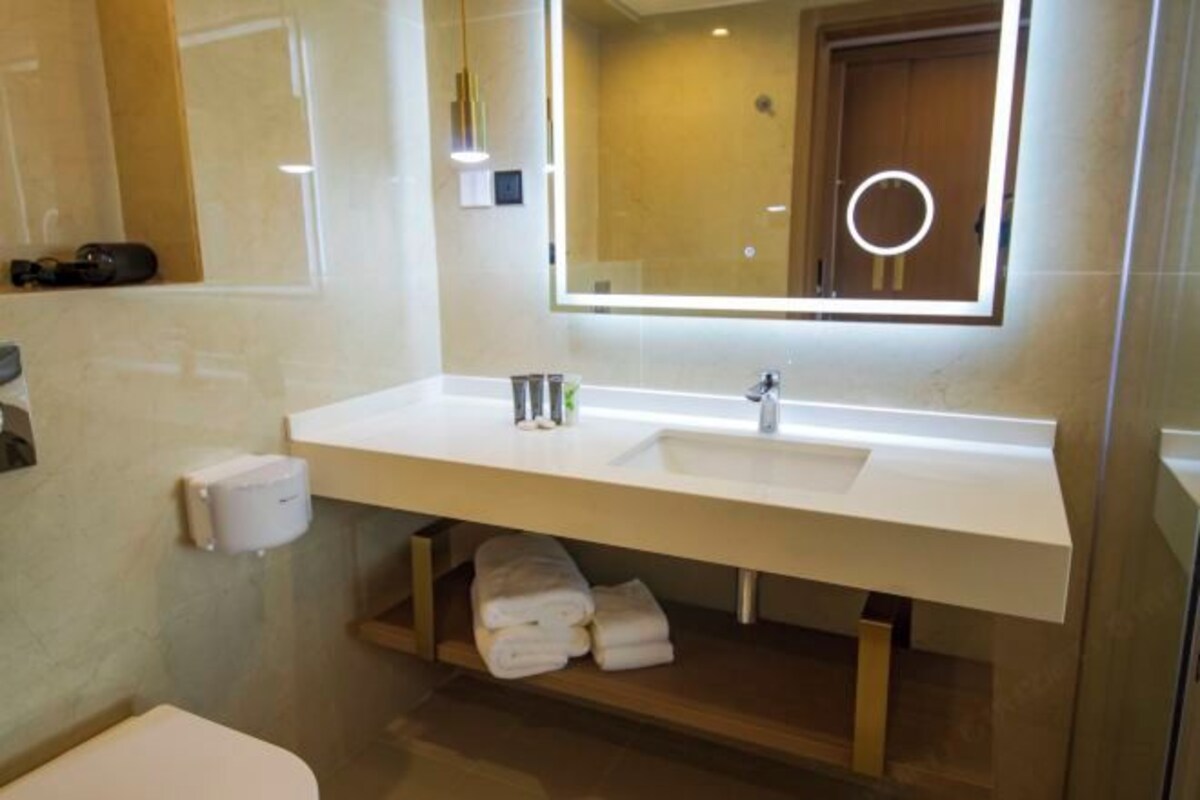 Une salle de bain moderne avec un lavabo blanc, un miroir doté d’un cadre lumineux, des toilettes et une étagère où sont pliées des serviettes.