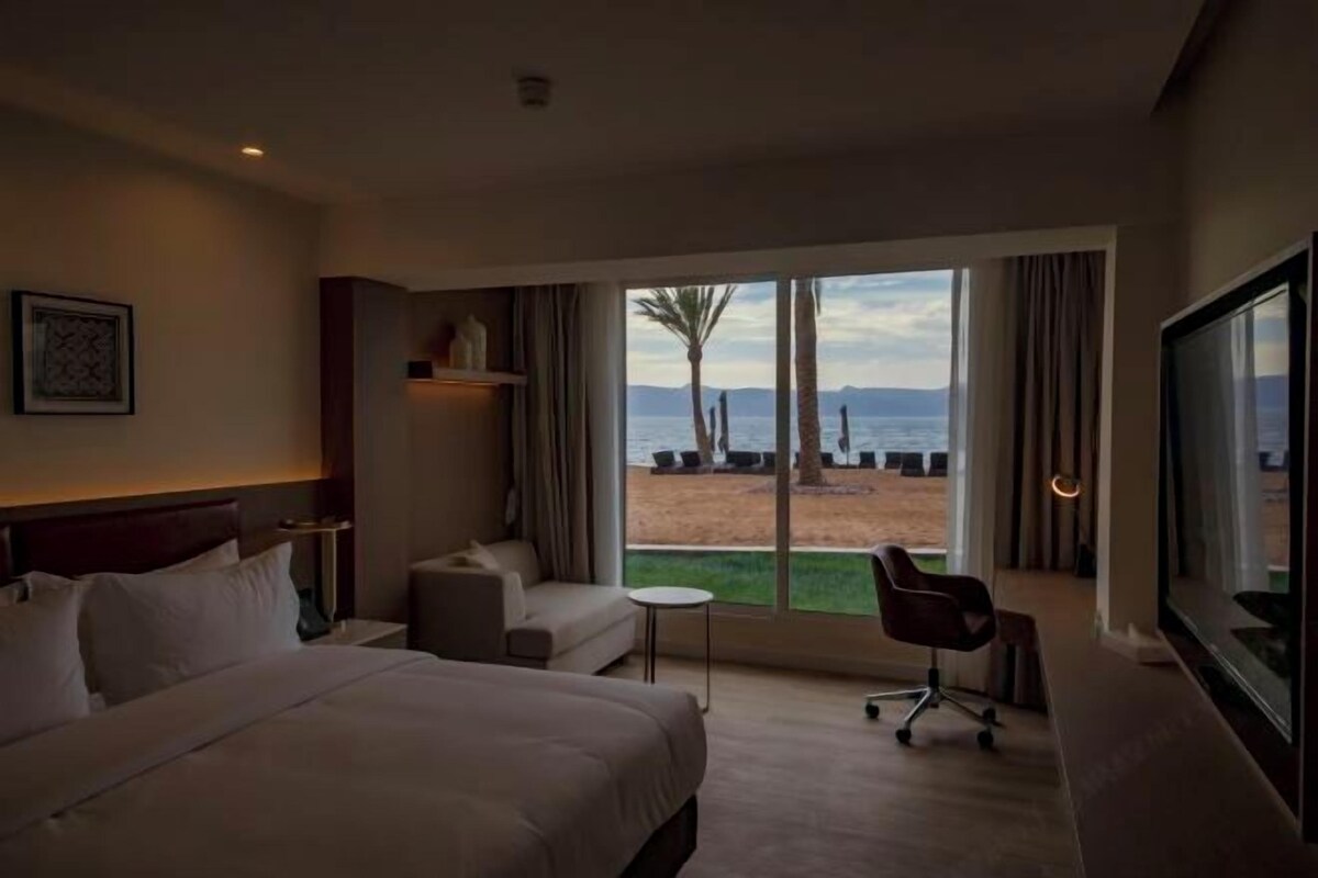 Ein Hotelzimmer mit einem großen Bett, einer kleinen Couch, einem Sessel und einem Fernseher. Ein großes Fenster bietet Blick auf einen Strand und Palmen.