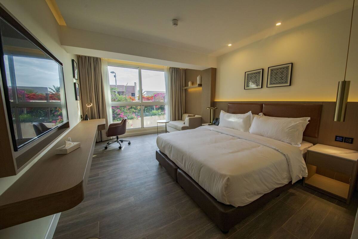 Une chambre d’hôtel avec un grand lit, un bureau, une chaise et une vue sur un balcon fleuri.