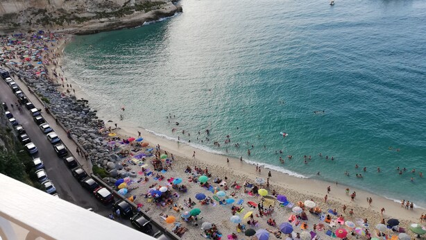 A coastal beach with a rocky cliff, a road with parked cars, and a clear view of the sea with boats. - Casa AuroraRosa (Tropea)