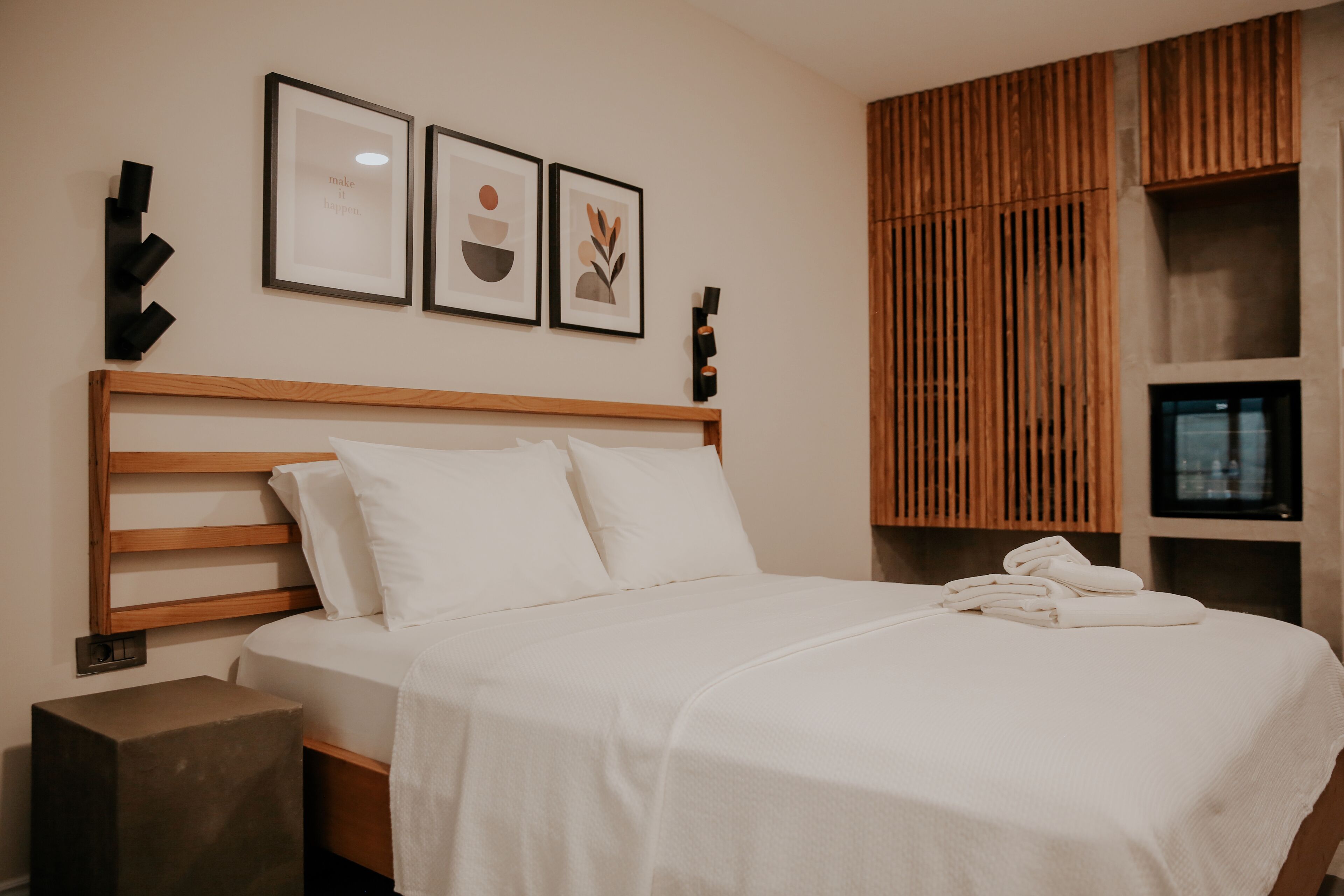 A neatly made bed with white linens, a wooden headboard, and a small bedside table with a lamp. There are framed artworks on the wall above the bed.