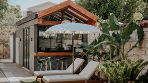 A modern outdoor bar area with two lounge chairs under a large umbrella, surrounded by lush greenery and a stone wall. - Paye Alaçatı (Cesme)