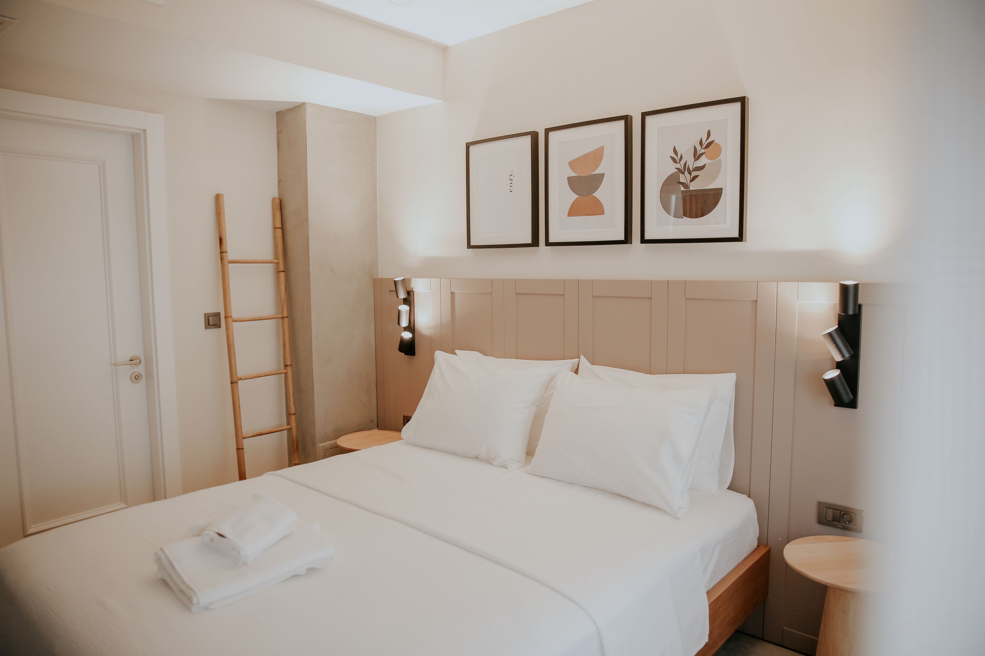 Un lit bien fait, avec du linge de lit blanc et une serviette pliée, une échelle en bois appuyée contre le mur, et trois œuvres d’art encadrées au-dessus du lit.