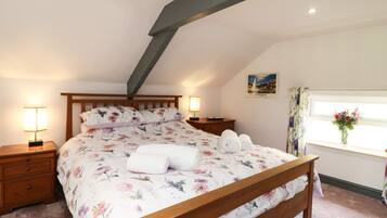 Un dormitorio con cama de madera, ropa de cama floral, una mesita de noche con lámpara, una ventana con cortinas y un cuadro en la pared.