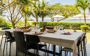 Mesa para comer al aire libre con sillas, platos, vasos y vista al mar.