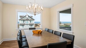 Ein Esszimmer mit großem Tisch, schwarzen Stühlen, einer Chandelierleuchtre und Blick auf ein Haus und den Strand.
